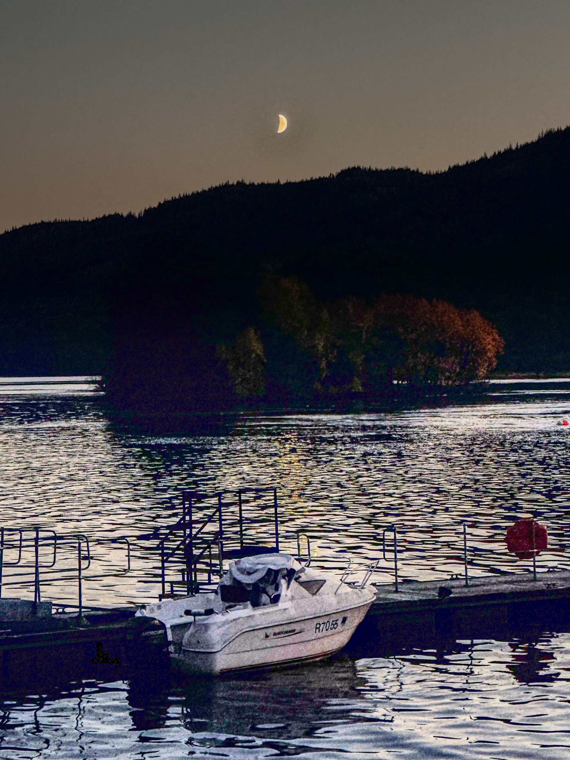 A waxing crescent just above the dark ridge at Inveruglas, with a boat moored at the jetty on Loch Lomond.